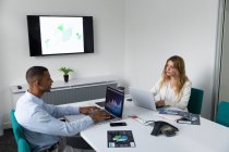 Side view of a young African American man and a young Caucasian woman sitting at opposite sides of a desk talking and using laptop computers in the modern office of a creative business — Stock Photo