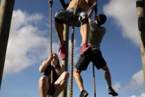 Vue en angle bas de deux jeunes femmes caucasiennes et d'un jeune homme caucasien escaladant des cordes dans un gymnase extérieur lors d'une séance d'entraînement de bootcamp — Photo de stock