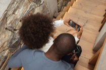 Visão traseira aérea de uma jovem mulher de raça mista e um jovem afro-americano tirando uma selfie com um smartphone sentado nas escadas em casa . — Fotografia de Stock