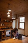 Повышенный вид молодой женщины смешанной расы с помощью ноутбука, сидящего у ее кухонного стола дома, ее смартфона и книги рядом с ней — стоковое фото