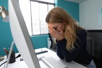 Vista lateral de una joven mujer caucásica sentada en un escritorio usando una computadora con la cabeza en las manos en la oficina moderna de un negocio creativo - foto de stock