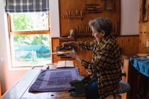 Seitenansicht einer älteren kaukasischen Geigenbauerin, die in ihrer Werkstatt den Körper einer Geige misst, im Hintergrund hängen Werkzeuge an der Wand — Stockfoto