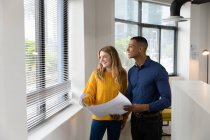 Front view of a young African American man and a young Caucasian woman holding plans and looking out of the window together standing in the modern office of a creative business — Stock Photo
