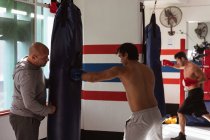 Seitenansicht eines jungen gemischten Boxers, der einen Boxsack schlägt, der von einem kaukasischen Trainer mittleren Alters gehalten wird, während im Hintergrund ein junger kaukasischer Boxer trainiert — Stockfoto