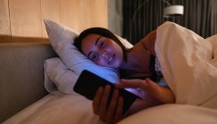 Visão frontal close-up de uma jovem morena caucasiana sorridente deitada de lado na cama usando um smartphone — Fotografia de Stock