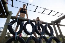 Vue de face de deux jeunes femmes caucasiennes escaladant un mur de pneus sur un cadre d'escalade dans une salle de gym extérieure lors d'une séance d'entraînement de bootcamp — Photo de stock