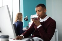 Frontansicht eines jungen afrikanisch-amerikanischen Mannes, der im modernen Büro eines kreativen Unternehmens an einem Schreibtisch sitzt und mit einem Smartphone spricht, während seine Kollegin im Hintergrund arbeitet — Stockfoto