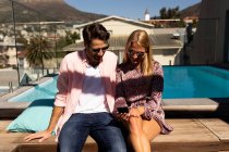 Vista frontal de una feliz pareja caucásica joven relajándose en vacaciones sentado junto a una piscina, utilizando un teléfono inteligente - foto de stock