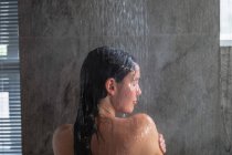Vista da vicino posteriore di una giovane donna bruna caucasica in piedi sotto una doccia che si lava i capelli, con la testa girata di lato in un bagno moderno — Foto stock