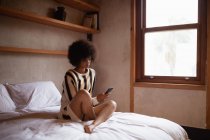 Vista lateral de una joven mujer de raza mixta usando un teléfono inteligente sentado en su cama en casa - foto de stock