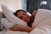 Ritratto da vicino di una giovane donna bruna caucasica sdraiata sul fianco nel letto sorridente alla telecamera — Foto stock