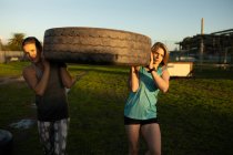 Vista frontal de duas jovens mulheres caucasianas carregando um pneu em um ginásio ao ar livre durante uma sessão de treinamento de bootcamp — Fotografia de Stock