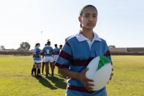Porträt einer jungen erwachsenen gemischten Rugbyspielerin, die auf einem Rugbyfeld steht und einen Rugbyball in der Hand hält und in die Kamera schaut, während ihre Teamkollegen im Hintergrund miteinander reden — Stockfoto