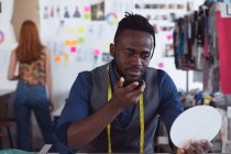 Vista frontal close-up de um jovem afro-americano estudante de moda masculina segurando e falando em um smartphone trabalhando em um design em um estúdio na faculdade de moda — Fotografia de Stock
