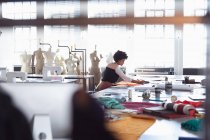 Vista laterale di una giovane studentessa di moda mista seduta a un tavolo che lavora su un design in uno studio del fashion college, con manichini sullo sfondo — Foto stock