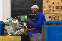 Зворотний вид молодого афроамериканського робітника сидячи і перевіряючи обладнання в машинобудівному цеху, з обладнанням і інструментами на задньому плані. — стокове фото