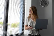 Front view close up of a young Caucasian woman standing holding a laptop computer with her head turned looking out of a window with Venetian blinds in the modern office of a creative business — Stock Photo