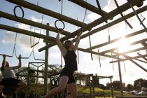 Side view of a young Caucasian woman hanging from rings on a climbing frame at an outdoor gym during a bootcamp training session, with a man climbing in the background — Stock Photo