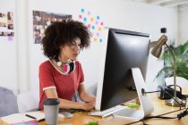 Vista lateral de uma jovem mulher de raça mista sentada em uma mesa digitando e olhando para uma tela de computador em um escritório criativo — Fotografia de Stock