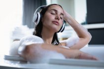 Вид спереди на молодую брюнетку, сидящую в пенной ванне в наушниках и слушающую музыку с закрытыми глазами — стоковое фото