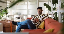 Вид спереди на молодого человека смешанной расы, сидящего на диване и использующего смартфон в креативном офисе — стоковое фото