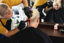 Visão traseira close-up de um cabeleireiro masculino caucasiano de meia idade e uma jovem mulher caucasiana com seu cabelo colorido vermelho brilhante em um salão de cabeleireiro, refletido em um espelho — Fotografia de Stock