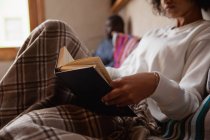 Вид сбоку на молодую женщину смешанной расы, сидящую на диване и читающую книгу дома, ее партнер, молодой афроамериканец, сидит на диване на заднем плане . — стоковое фото