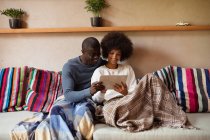 Vista frontale da vicino di una giovane donna di razza mista e un giovane uomo afroamericano che guarda un tablet e parla seduti insieme su un divano a casa
. — Foto stock