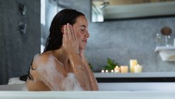 Side view close up of a young Caucasian brunette woman sitting in a bath with lit candles on the side, washing her face with her eyes closed — Stock Photo