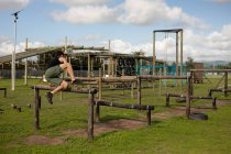 Vista lateral de um jovem caucasiano saltando sobre um obstáculo em um ginásio ao ar livre durante uma sessão de treinamento de bootcamp — Fotografia de Stock