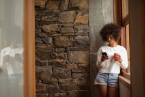 Frontansicht einer jungen Frau mit gemischter Rasse, die am Fenster steht, ein Smartphone benutzt und eine Tasse Kaffee in der Hand hält — Stockfoto