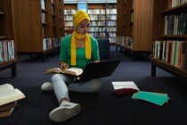Vista frontal close up de uma jovem estudante asiática vestindo um hijab segurando um livro, usando um computador portátil e estudando em uma biblioteca — Fotografia de Stock