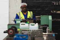Vista frontal de perto de um jovem trabalhador da fábrica afro-americano sentado e operando equipamentos na loja de máquinas em uma fábrica de processamento, com equipamentos e ferramentas em primeiro plano — Fotografia de Stock