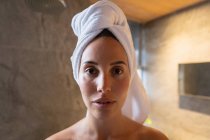 Porträt einer jungen kaukasischen brünetten Frau mit in ein Handtuch gewickelten Haaren, die in einem modernen Badezimmer direkt in die Kamera blickt — Stockfoto