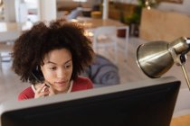 Nahaufnahme einer jungen Frau mit gemischter Rasse, die an einem Schreibtisch sitzt, ein Headset trägt und in einem Kreativbüro auf einen Computerbildschirm blickt — Stockfoto