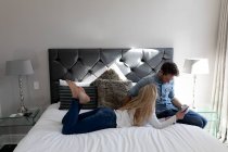 Vista lateral de um jovem casal caucasiano feliz relaxando juntos de férias em um quarto de hotel sentado e deitado em uma cama usando um smartphone — Fotografia de Stock