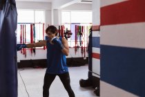 Vue latérale d'un jeune boxeur masculin métis dans un gymnase de boxe frappant un punchbag — Photo de stock