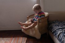 Vista laterale di un bambino caucasico seduto su una sedia e con uno smartphone in mano, a piedi nudi — Foto stock