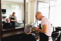 Vue latérale d'un coiffeur caucasien d'âge moyen et d'une jeune femme caucasienne se faisant couper les cheveux dans un salon de coiffure, reflétée dans un miroir — Photo de stock