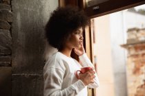 Seitenansicht einer jungen Frau mit gemischter Rasse, die am Fenster steht und mit einer Tasse Kaffee nach draußen schaut — Stockfoto
