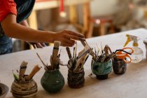 Gros plan de la main d'une potière debout et sélection d'un outil à partir de pots d'outils sur une table de travail dans un atelier de poterie — Photo de stock