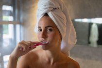 Portrait close up of a young Caucasian woman with her hair wrapped in a towel brushing her teeth and looking to camera in a modern bathroom — Stock Photo