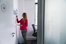 Side view close up of a young Caucasian woman standing holding a tablet computer and reading colored sticky notes on a wall in the modern office of a creative business — Stock Photo