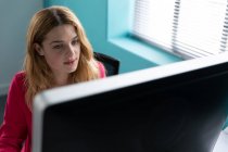 Vista frontal fechar uma jovem mulher caucasiana sentada em uma mesa por uma janela usando um computador no escritório moderno de um negócio criativo — Fotografia de Stock