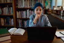 Передній вид на молоду азіатську студентку, одягнену в тюрбан з допомогою ноутбука і навчаючись у бібліотеці. — стокове фото