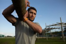 Nahaufnahme eines jungen kaukasischen Mannes, der während eines Bootcamp-Trainings einen Holzklotz in einem Outdoor-Fitnessstudio trägt — Stockfoto