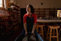 Vue de face d'une jeune potière caucasienne assise avec un pot sur une roue de potiers dans un atelier de poterie, et regardant vers la fenêtre — Photo de stock