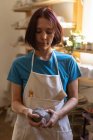 Вид спереди на молодую кавказскую гончарку в фартуке, смотрящую на кусок глины, который она держит в руках, и позирующую в керамической мастерской — стоковое фото
