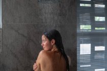 Вид сзади на молодую кавказскую брюнетку, стоящую под душем, моющую голову, отвернутую в сторону в современной ванной — стоковое фото