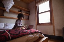 Вид збоку на молоду змішану жінку, яка використовує ноутбук, сидячи на своєму ліжку вдома — стокове фото